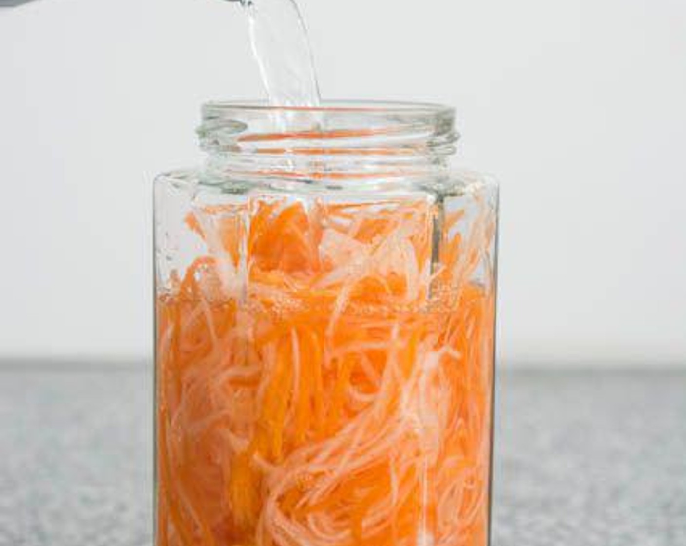 Schritt 1 Für das eingelegte Gemüse: Gestiftelte Karotten (150 g) und gestiftelten Rettich (150 g) in ein Glas geben. Das Glas mit Essig, mild (240 ml), Wasser (180 ml) und Zucker (4 Esslöffel) auffüllen.